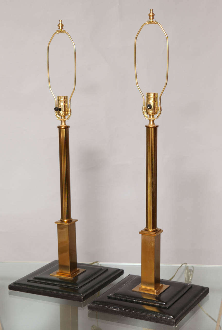 A pair of French Jacques Adnet style table lamps, the reeded brass stem resting on square pedestal above stepped leather bases, with hand-stitched detail.