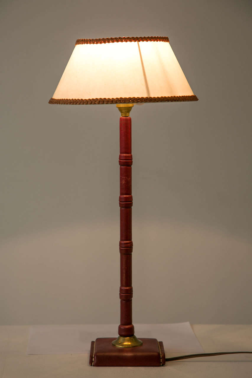 Table lamp by Jacques Adnet (1900-1984) entirely covered with saddled stitched red leather. Tubular trunk and two sided openwork base jointed by a gold metal strapping. Lamp shade with flared sides. Wired for continental Europe use.