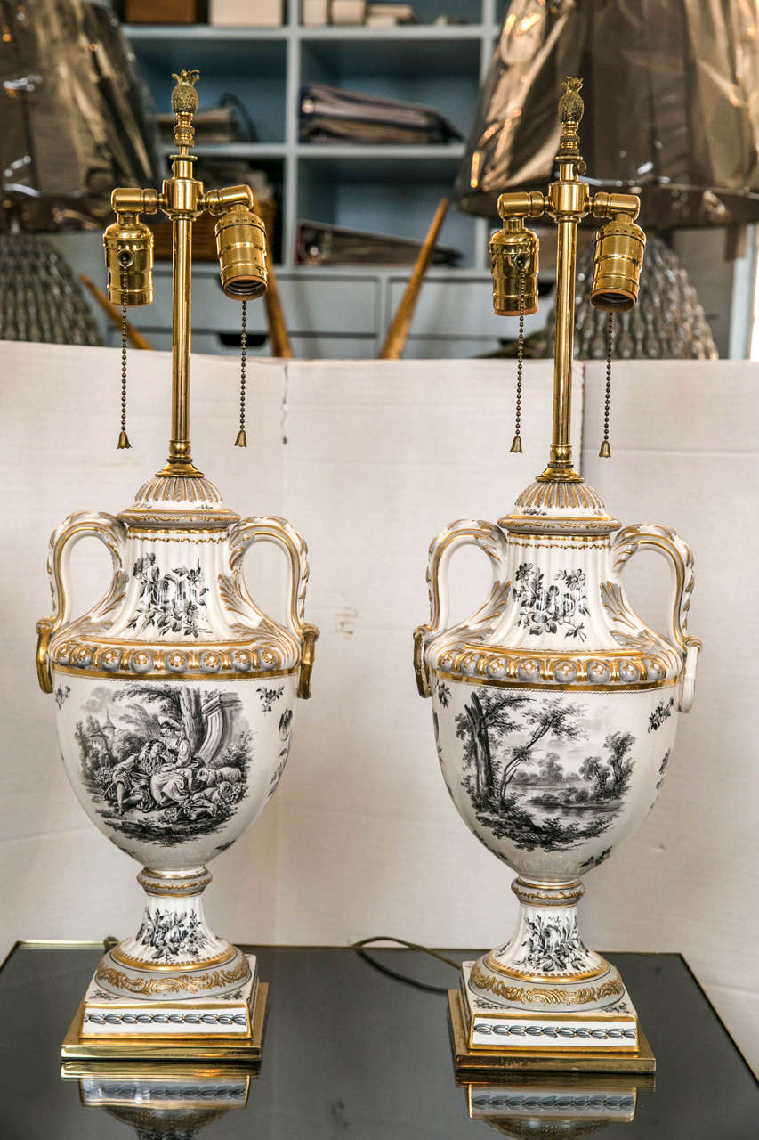 A pair of porcelain urn vases with a black and white toile style decoration and gilded embellishment. The square bases are brass and all hardware is brass. No shades are included.