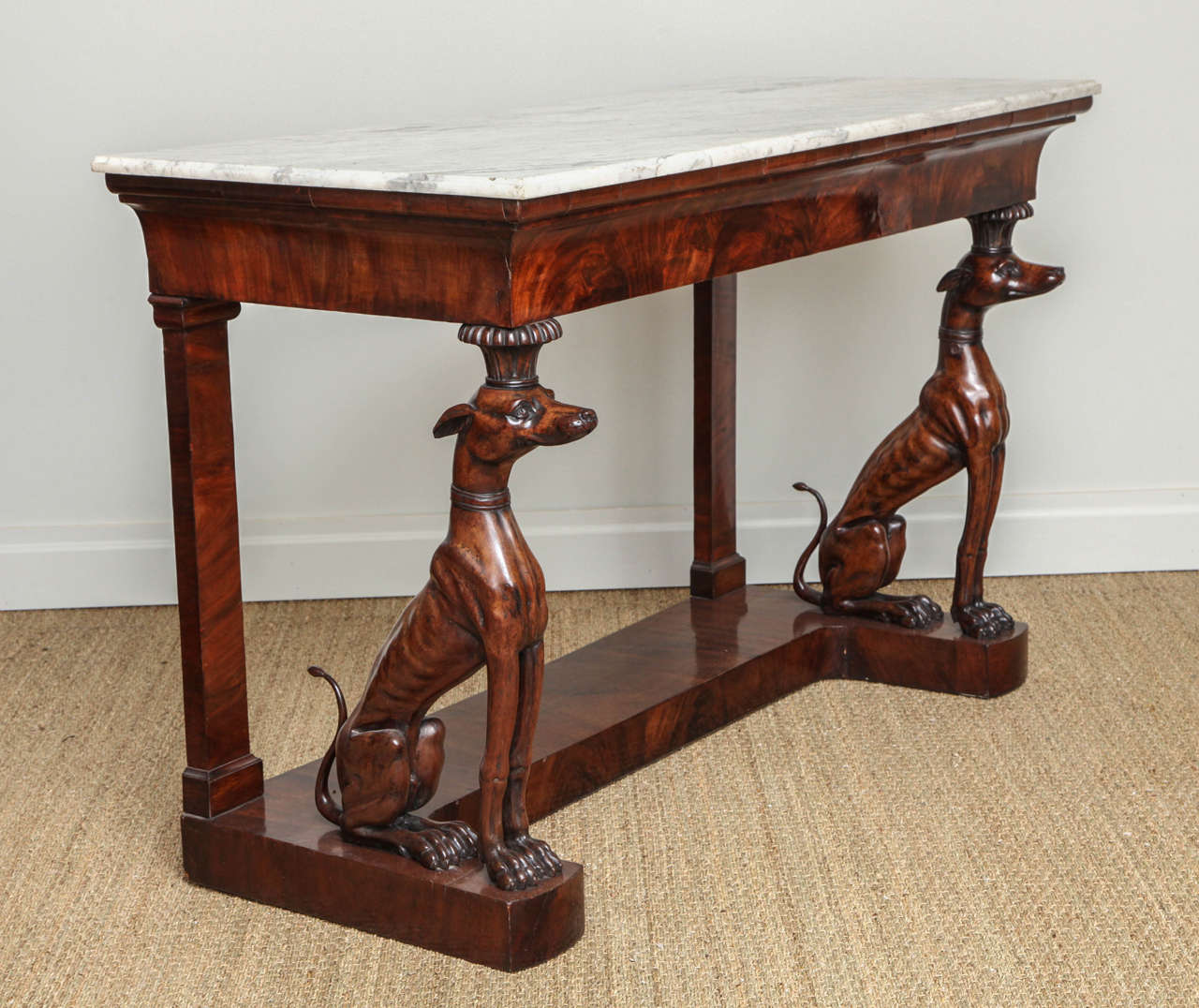 An impressive Italian marble-top mahogany console table, the original Carrara marble top with cove edge over molded crotch grained frieze, supported by finely carved and whimsical whippet supports on a shaped plinth base, the whole with good patina