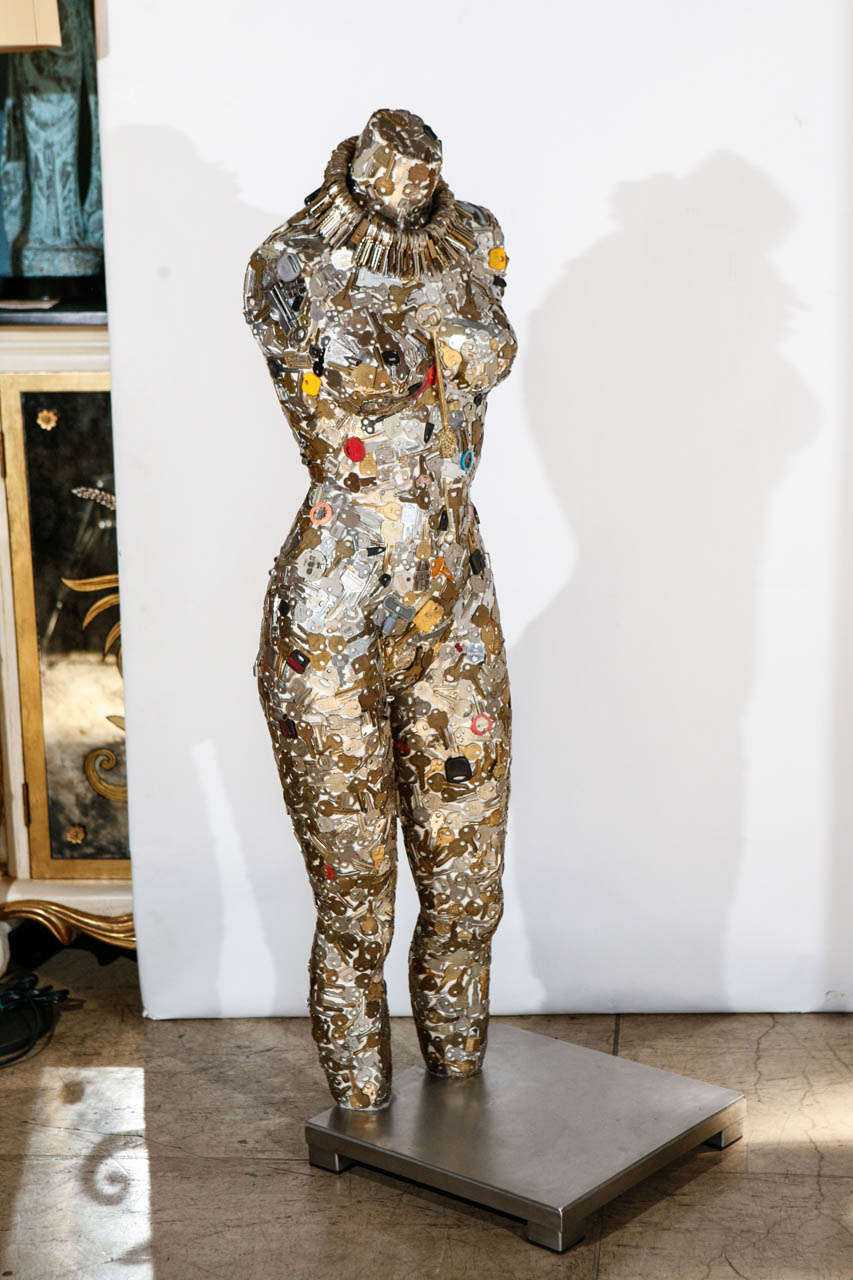 A tall form woman mannequin sculpture mounted on a brushed steel base. Sculpture is covered with vintage keys. Sculpture is accessorized with a choker also made of keys.