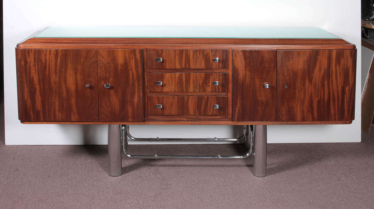 A French Modern credenza. The central frieze of three drawers, double side doors bearing radius corners with original polished hardware, and a gently sloping bull nose apron inset with a frosted glass top, is gracefully mounted, appearing as if