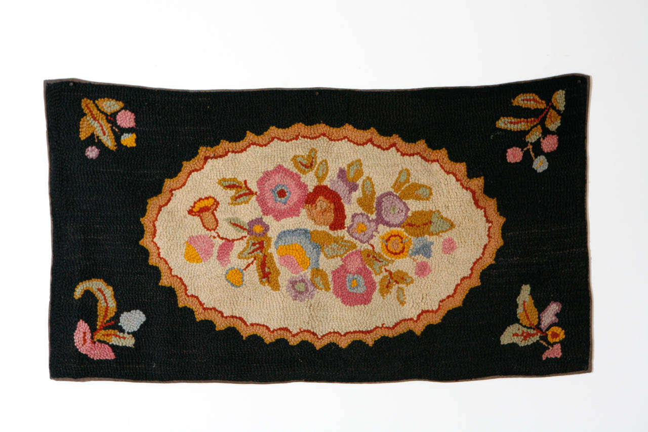 American hand hooked wool rug of brightly colored floral bouquet on oval, pale gray background, framed in black wool surround, with small floral motifs in each of the four corners.