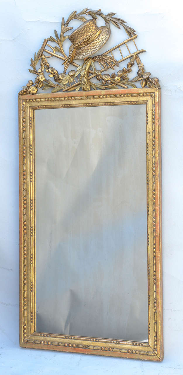 Gitlwood mirror, having a rectangular looking glass of somewhat spotted/distressed mirrorplate, in molded frame decorated by bead-and-bar motif, surmounted by elaborately carved pierced pediment consisting of straw hat, rake, ladder and festooned