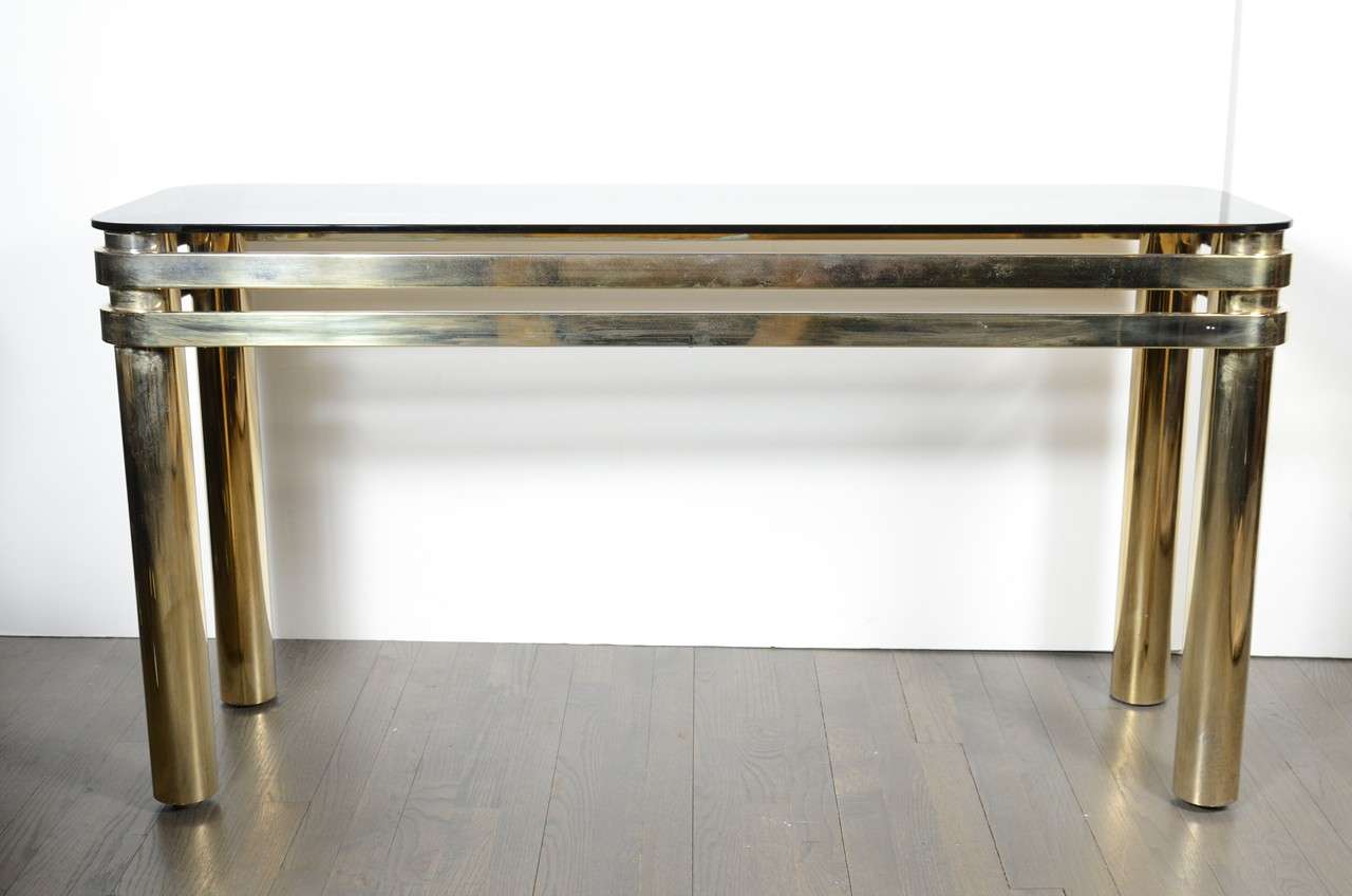 This fine Mid Century Modern brass console table was realized in the United States circa 1970. It features features double banded supports and smoked glass top. With its bold forms and luxe materials, it embodies the characteristics that enthusiasts