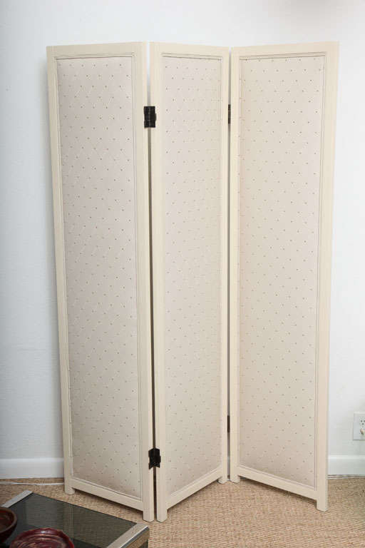 Trifold room divider fashioned of bone colored lacquered wood. Front and back panels are cushioned and upholstered in diamond pattern faux leather with gold thread and finished with thin double welting. Bronze hinges allow full rotation of each