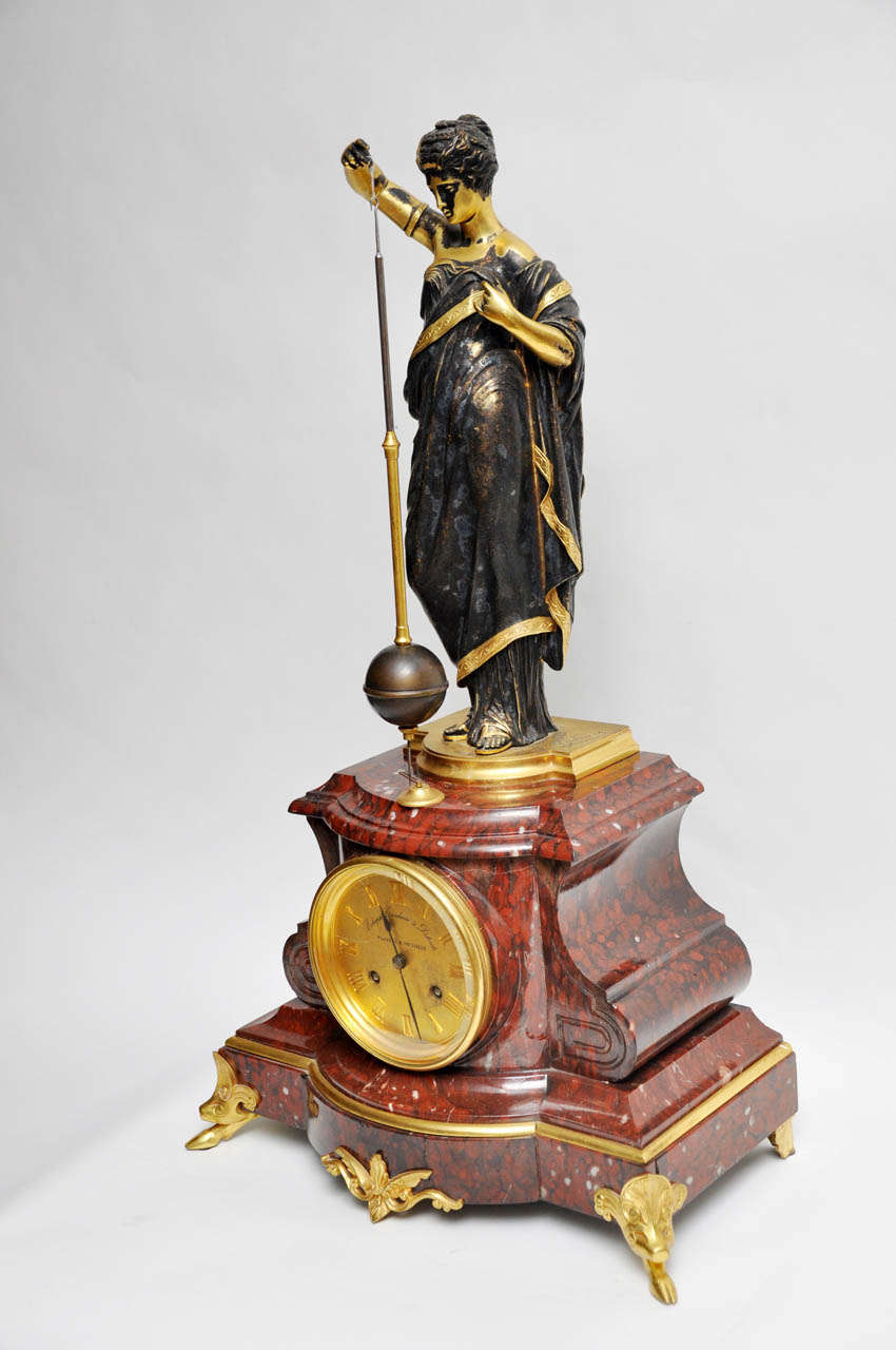 A gold bronze statue of a Grecian Woman, supported by a royal rouge marble clock. Gold bronze accents woman's dress, feet, arms and neck, dress is in natural patina bronze. Suspended from the statue's right arm is the pendulum having a brass and