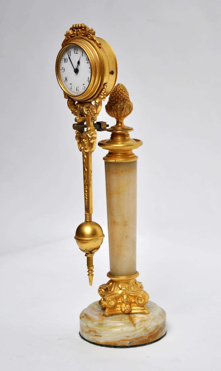 Rare French, gold bronze swinging arm clock mounted on an onyx column with gold bronze acorn finial, capitol and scrolled footed base, sitting on an onyx platform. Elaborate gold bronze surrounds the suspended, porcelain dial having black Arabic