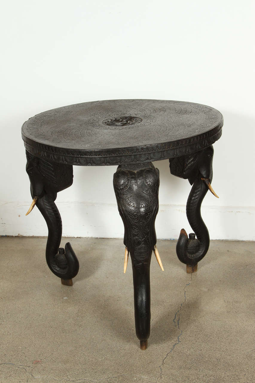 Intricately and finely hand-carved ebonized wood round Colonial Anglo Indian table. Three carved elephant heads with bone tusks holding jars of oil. 
These finely carved late 19th century tables are getting very hard to find.
All bone tusks are