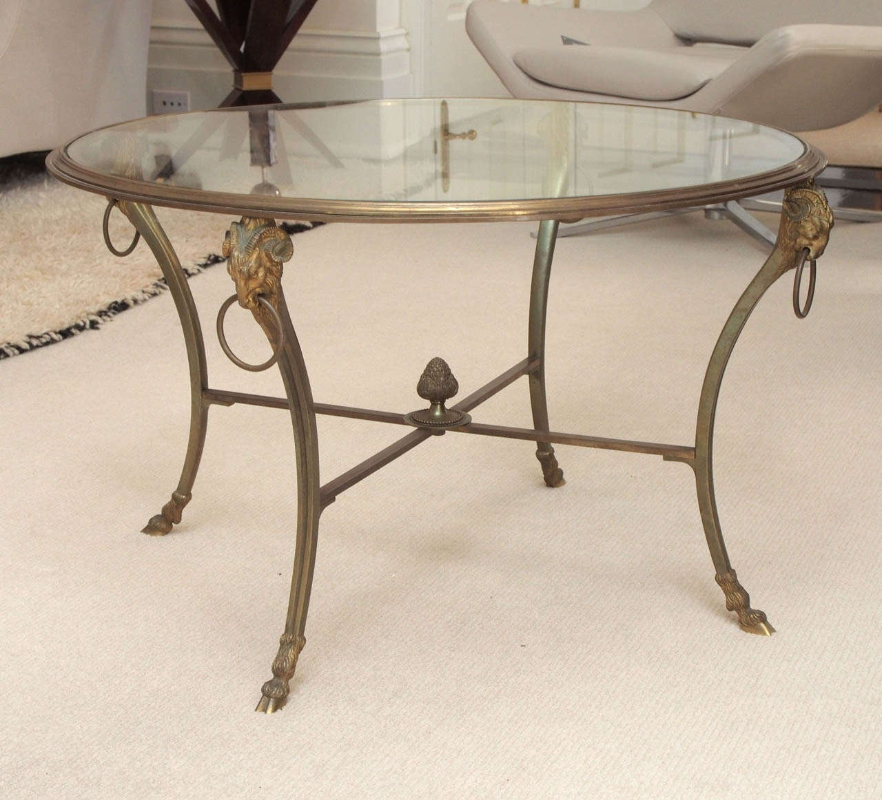 Low circular table in gilt bronze; elegantly detailed with hoof feet, ram heads and acorn finial at the stretchers; clear glass plateau