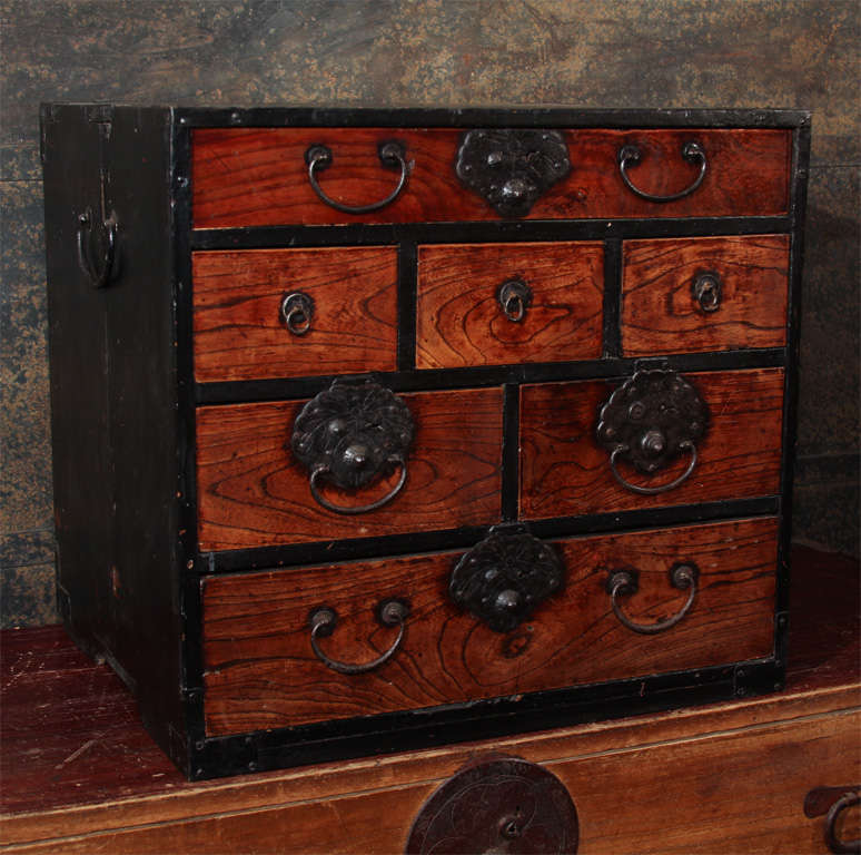 A small Japanese chest of drawers. The tansu with seven keyaki wood drawers of fine color and patina of various sizes housed within a sugi, cryptomeria case. The chest with iron fittings and hardware, including lock plates of paulownia, kiri leaf