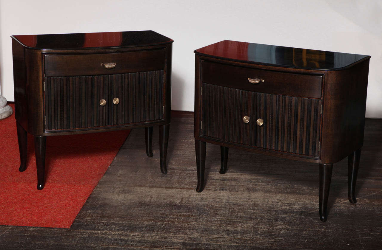 Pair of low bedside tables with ample storage on tall, tapering legs.  Dark stained wood, with polished brass pulls and ribbed detailing to door fronts.  Practical and great looking.