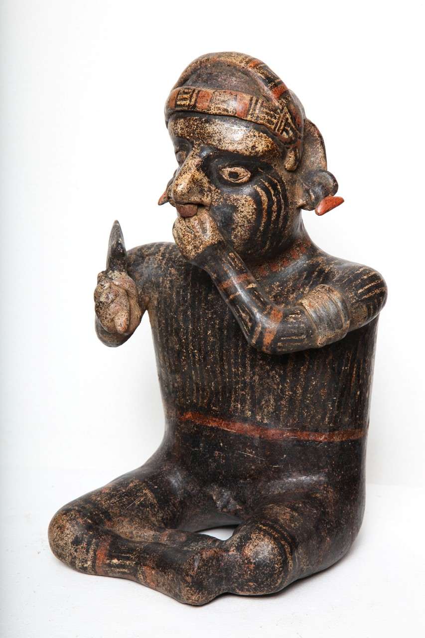 A well decorated sculpture of excellent form, a shaman or priest,  seated cross legged and holding a mace, he has pleasant facial features, ear clips and cap headdress and gestures one hand to his mouth. The surface is painted with the most amazing