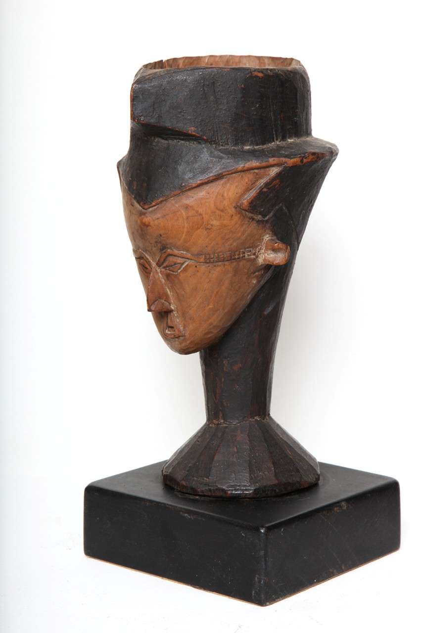 Carved palm wine cup rising from a circular base, the angular face with naturalistic slender facial features, compact pronounced ears and scarification at the sides of the eyes, and wearing a triangular cap, extending from hair. Painted two tone,