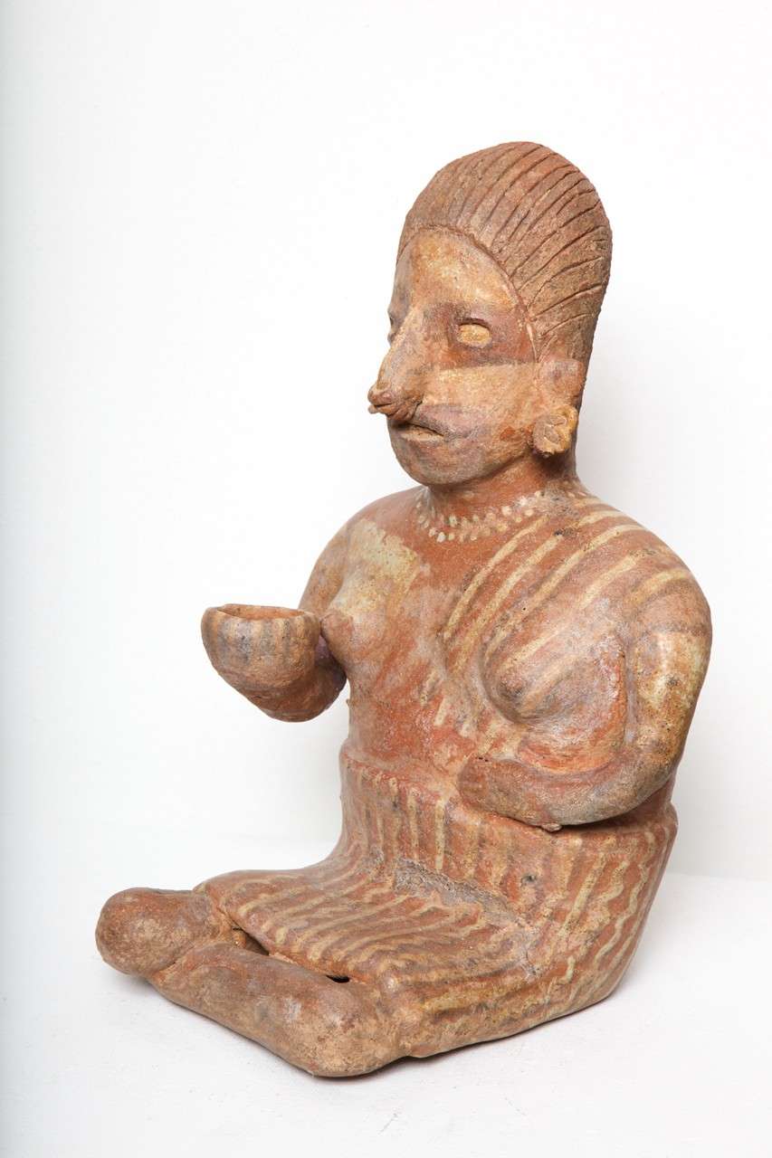 A unique modeled and painted figural effigy; she is seated side legged and holding a small vessel in her right hand.  She is adorned with earspools, nose clip, painted necklace, and she is wearing stripped pattern polychrome garments. 

Overall