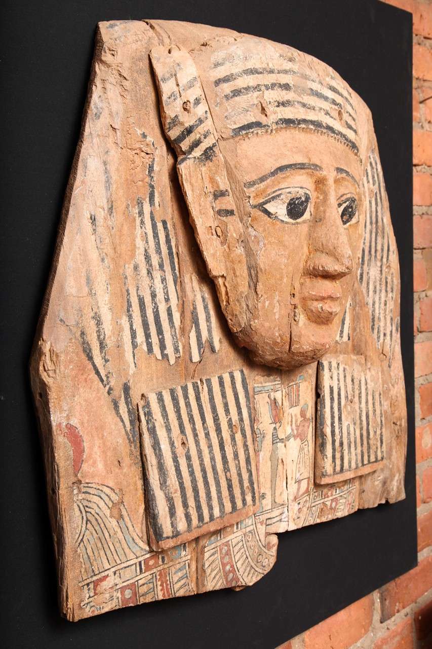 The mask with polychrome painted gesso details, shown wearing a striped black and white wig, the face carved with exaggerated features, painted between the lapels with the deceased, standing before Osiris, with an udjat eye below and remains of a