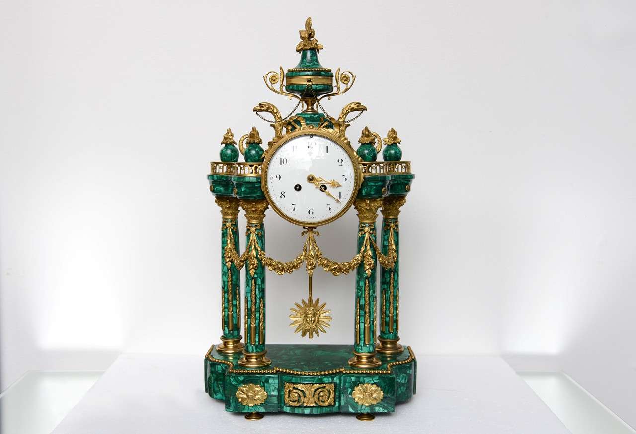 Neo-classic style ormolu and malachite mantel clock. Clock has an enamel face with Arabic numerals mounted on a structure in the form of a temple with four corinthian columns hung with floral swags, sur mounted by an eagle and urn finial. The