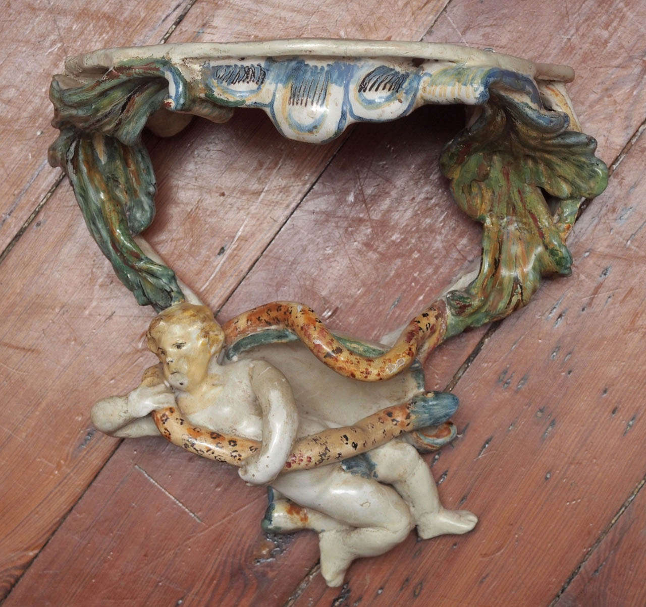 A small ornamental tin glazed ceramic wall bracket, the top supported by green foliate arms joined at the bottom by a nude figure of an infant holding a snake, possibly representing the infant Hercules.  This piece has areas of restoration,
