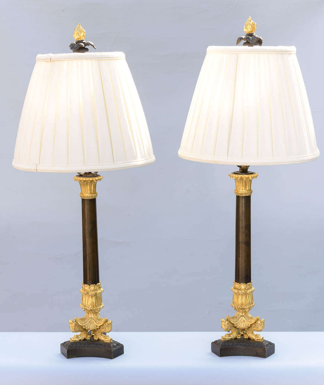Pair of lamps, having patinated bronze column with dore bronze acanthus capital, raised on tripartite base of dolphins, finished with period gilt bronze finial. Shown with pleated shades.