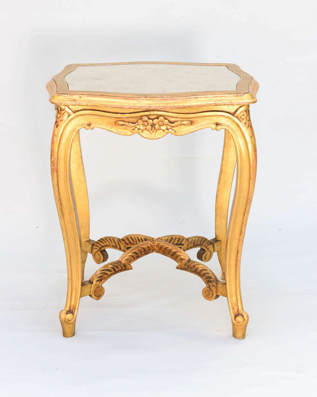 Carved giltwood accent table, having a distressed mirrored top inset in frame, apron with floral details, raised on cabriole legs connected by X-stretcher.
