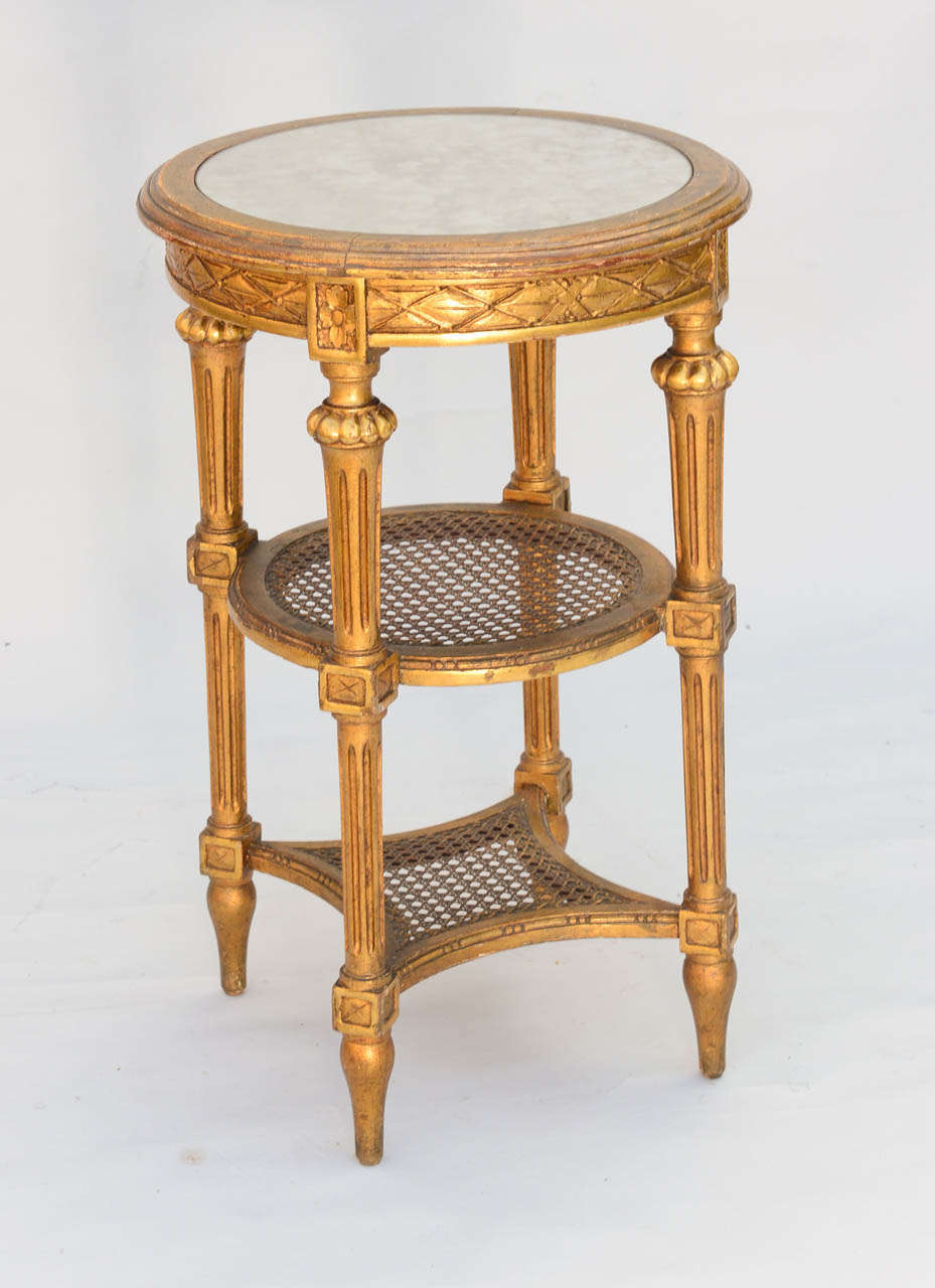 Round candlestand table or etagere, having mirrored top inset in giltwood frame; apron carved with crosshatch designs, raised on round fluted legs connected by caned stretchers, one round, the second with concave sides.
