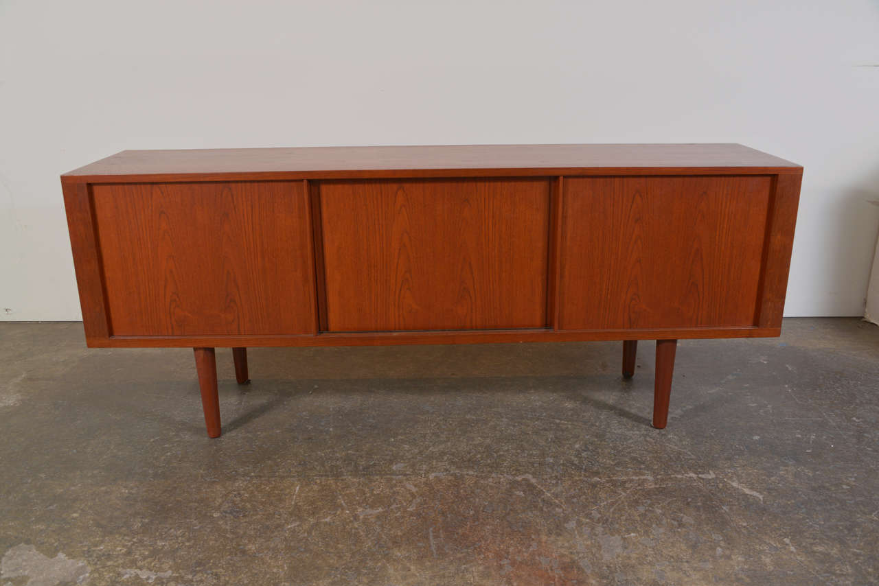 Solid teak wood constructed credenza designed Hans Wegner and manufactured by cabinetmaker of note, Ry Mobler. This simple box on legs features three sliding doors that conceal an oak lined interior. It has five shallow drawers behind the center