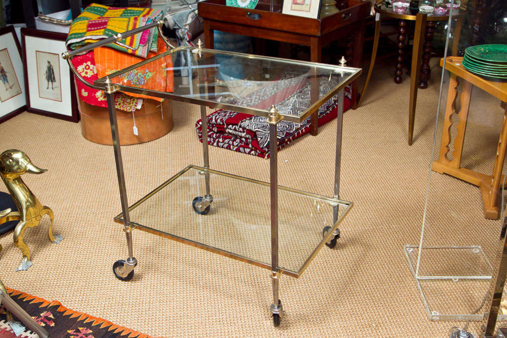 A two level brass bar cart of steel and brass. Inset with clear glass shelves, on large casters.