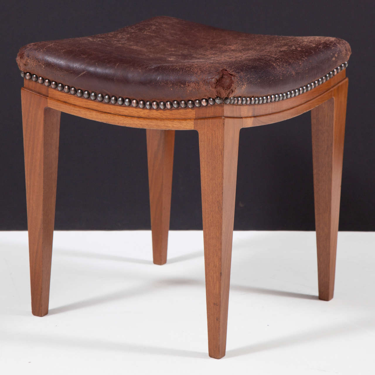 A single mahogany and leather upholstered stool, Circa 1940s, with a square leather top and french nailheads, channeled and curved frieze raised on square tapered legs.