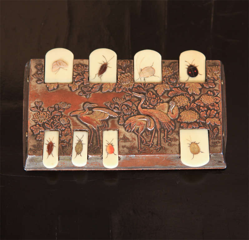 A fine Japanese silver plate and ivory whist marker. The surface of the case decorated in low relief with a garden scene of four cranes amid flowering peonies. The counter with eight hinged ivory tabs, each decorated with an insect of inlaid stone