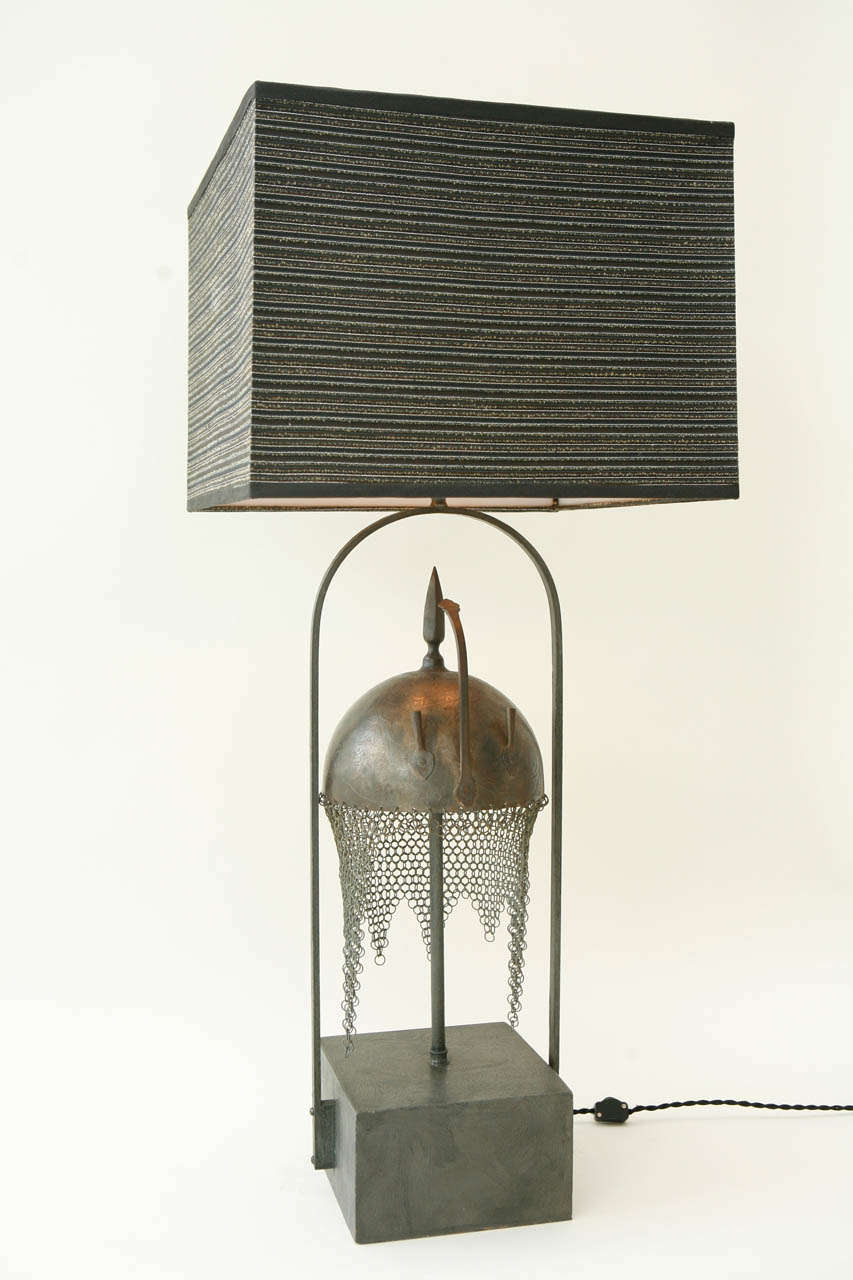 A dramatic armature table lamp with a textured painted wood base displaying a Persian Kulah Khud helmet and a black silk shade overlaid with a Lurex casement. The iron helmet is heavily engraved with figures and Arabic script and has a chain mail
