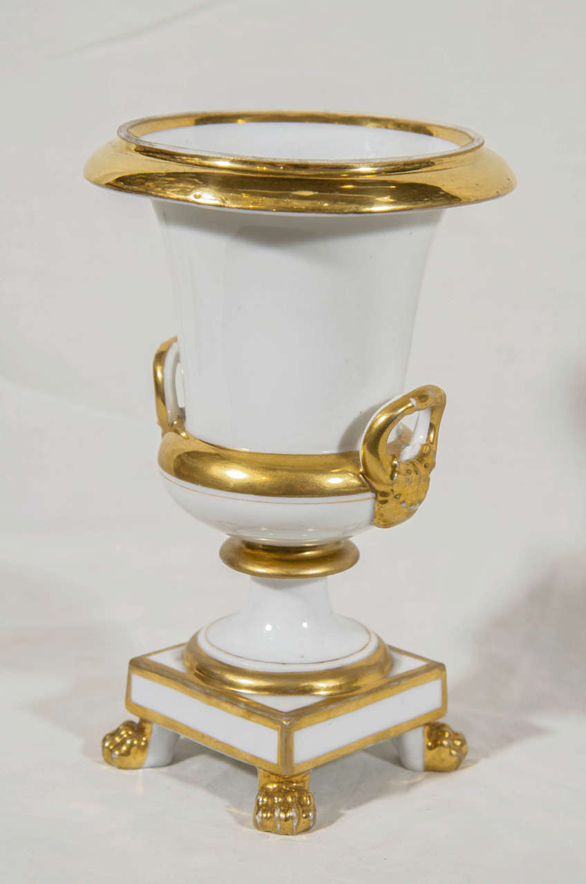 A pair of small Paris porcelain neoclassical white and gilt urns raised on square bases with lion's paw feet.
