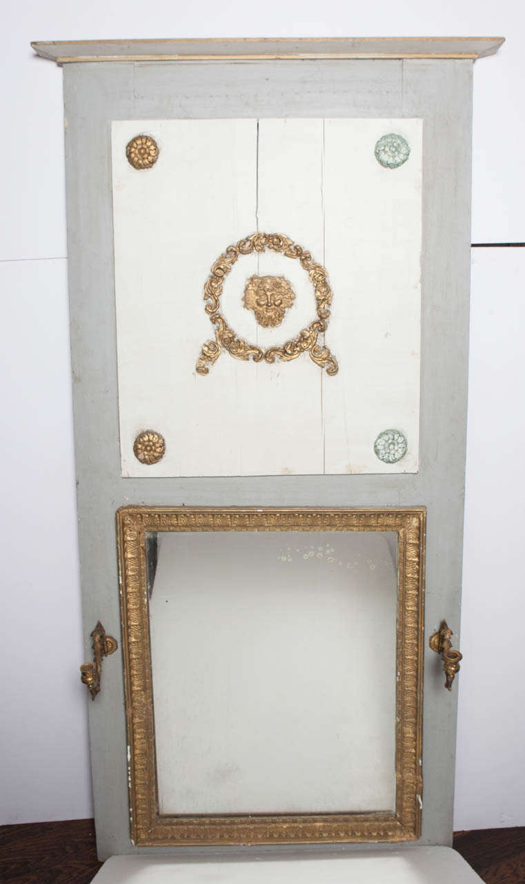 Gray and white painted wood trumeau/mirror with later additions.  Gold painted trim made of plaster.  Brass candle holders.  Top panel has an applied brass relief head.