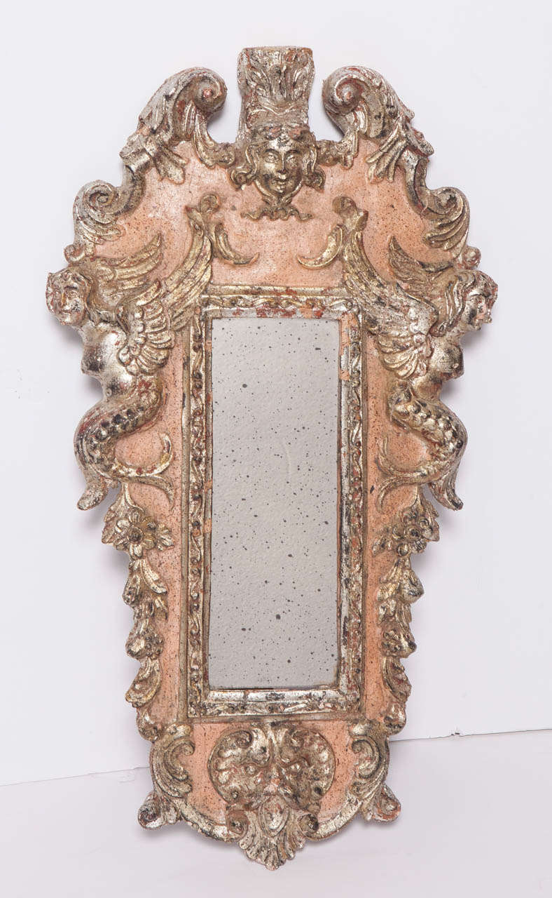 Painted petite Italian mirror has silver leaf detail on plaster and wood.  Mirror frame depicts two winged mermaids on each side, an angel at the top and appears to depict the Mouth of Truth at the bottom.  Replaced antiqued mirror.  Refreshed paint.