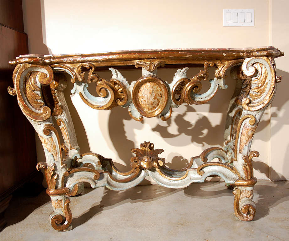A stunning, hand-carved and painted, parcel-gilt console table featuring a pierced apron above scroll feet and X-stretcher crowned by a relief-carved medallion. The whole surmounted by a rouge marble top.