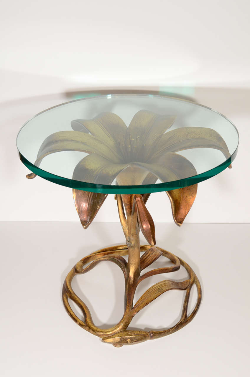 Stylized lily table with patinated gilt finish over metal bases and circular glass tops. The table has an Art Nouveau inspired design and features a base comprised of lily stems, leaves, and partially opened lillies, as well as a large sculpted lily