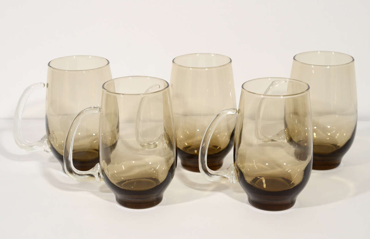 Set of six mid-century barware glasses or beer mugs in blown glass with smokey grey-brown color. Feature stylized clear glass handles with modernist horn design.