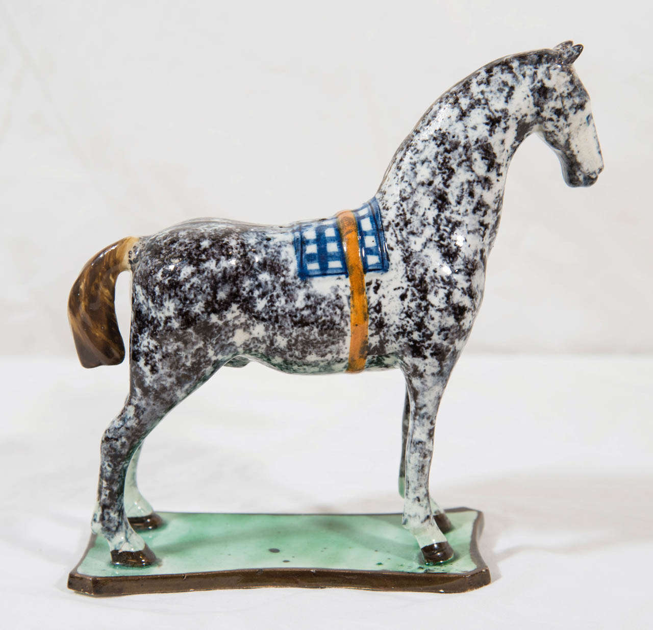 A dapple gray pearlware horse with a cropped tail standing on a flat green glazed slab base. A blue and white checkered saddle blanket rests on the horse's back held on by an orange leather strap.
 These horses were originally made to be gifts for