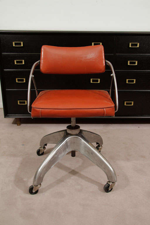 A machine age desk chair with a sturdy brushed steel base and tomato red vinyl cushions. The chair swivels on its base and the back cushion pivots for comfort. The piece is a charming example of industrial style.