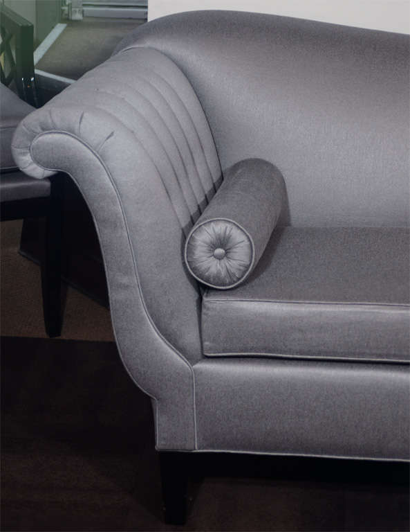 Realized in the United States circa 1945, this Hollywood Art Deco sofa embodies the understated elegance of a bygone era. It features a sinuously curved design; scrolled arms; tufted sides; and cylindrical bolsters with button detailing and pleated