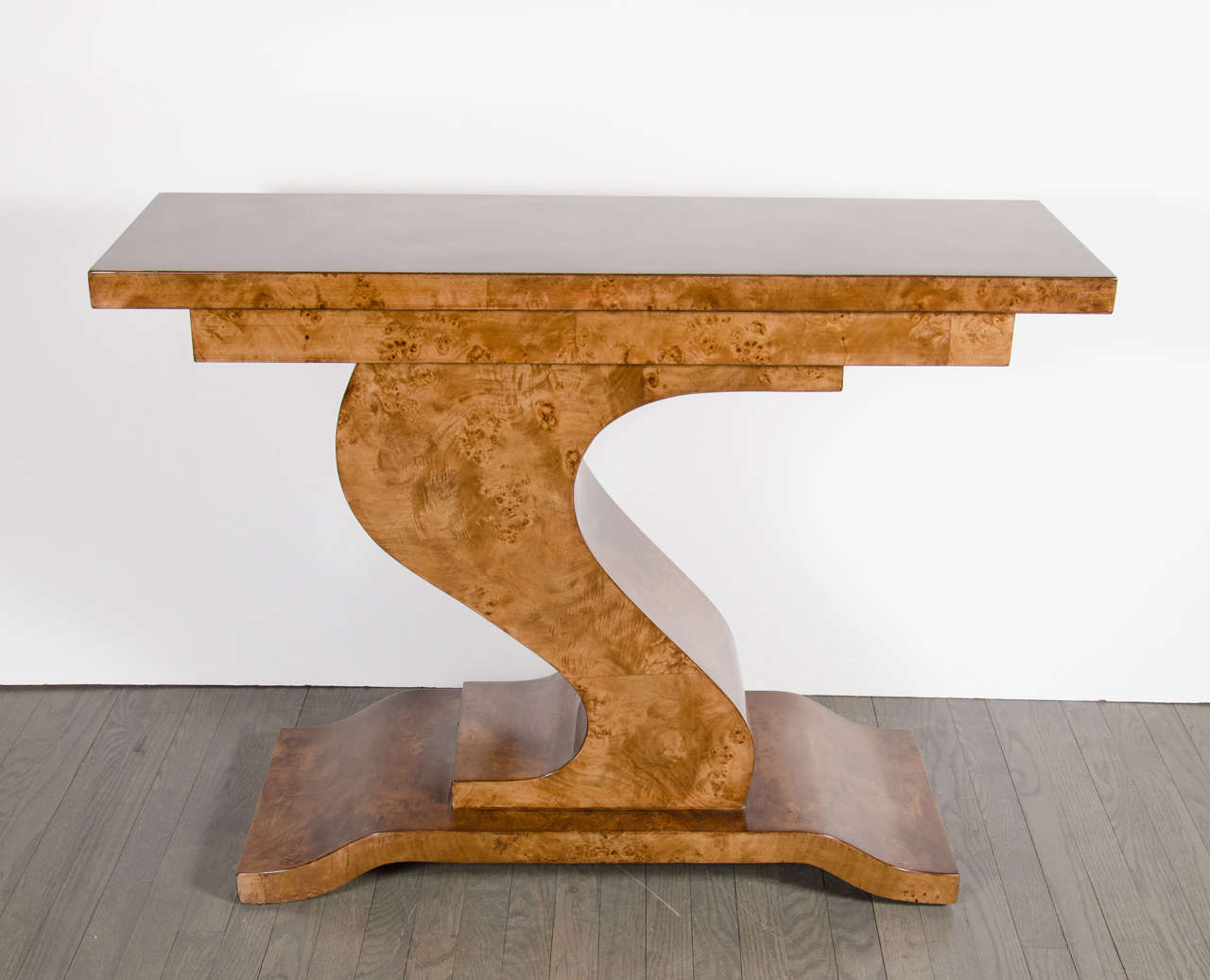 Pair of Mid-Century Modernist console tables in the manner of Karl Springer featured in an asymmetrical fluid design. This pair of exquisite consoles are showcased in their stunning book-matched carpathian elm that rests atop a raised plinth base
