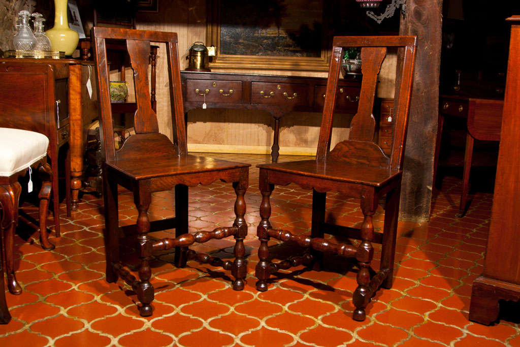 A fine, early pair of Georgian oak hall chairs with turned legs and stretchers, and canted splat backs.  These chairs date to the very beginning of the reign of George I and, as such,  bear more characteristics of Queen Anne (George I's predecessor)