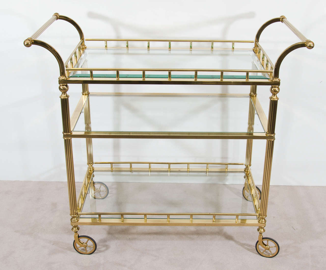 A vintage double handle brass bar cart with three tiers of glass shelves. The top shelf has beveled glass.