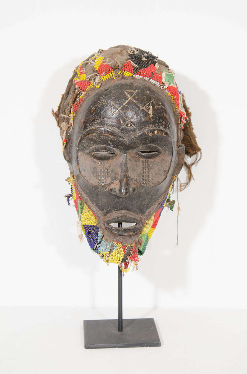 A tribal ritual wooden mask in the style of the Chokwe tribe of the Democratic Republic of Congo. Decorated with a cloth beaded multicolored headdress and depicting facial scarification patterns and cruciform markings on the forehead. Known for