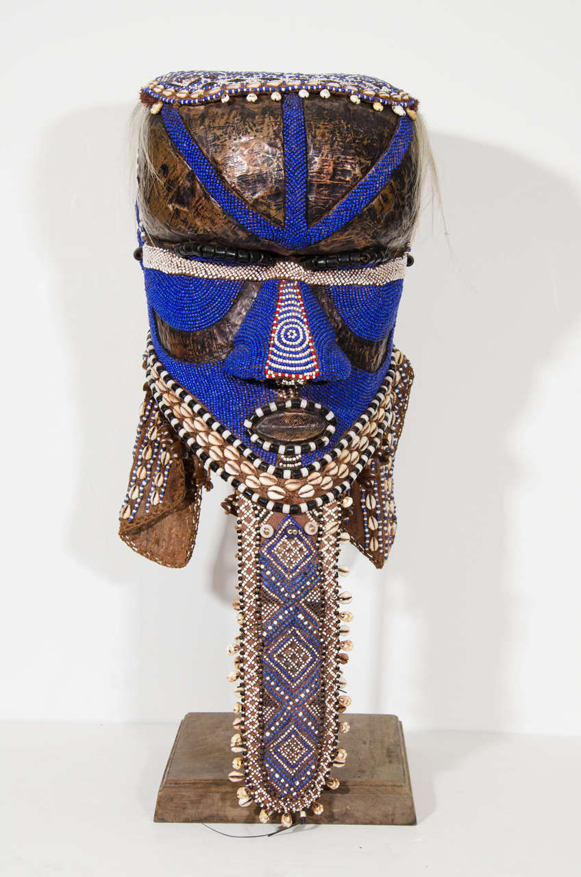 A 21st Century Kuba mask or helmet from the Kuba kingdom in the Democratic Republic of the Congo. The mask is embellished with bright blue and geometric designs in a variety of materials: beads, shell, cloth, hair, and copper sheet. The mask is worn