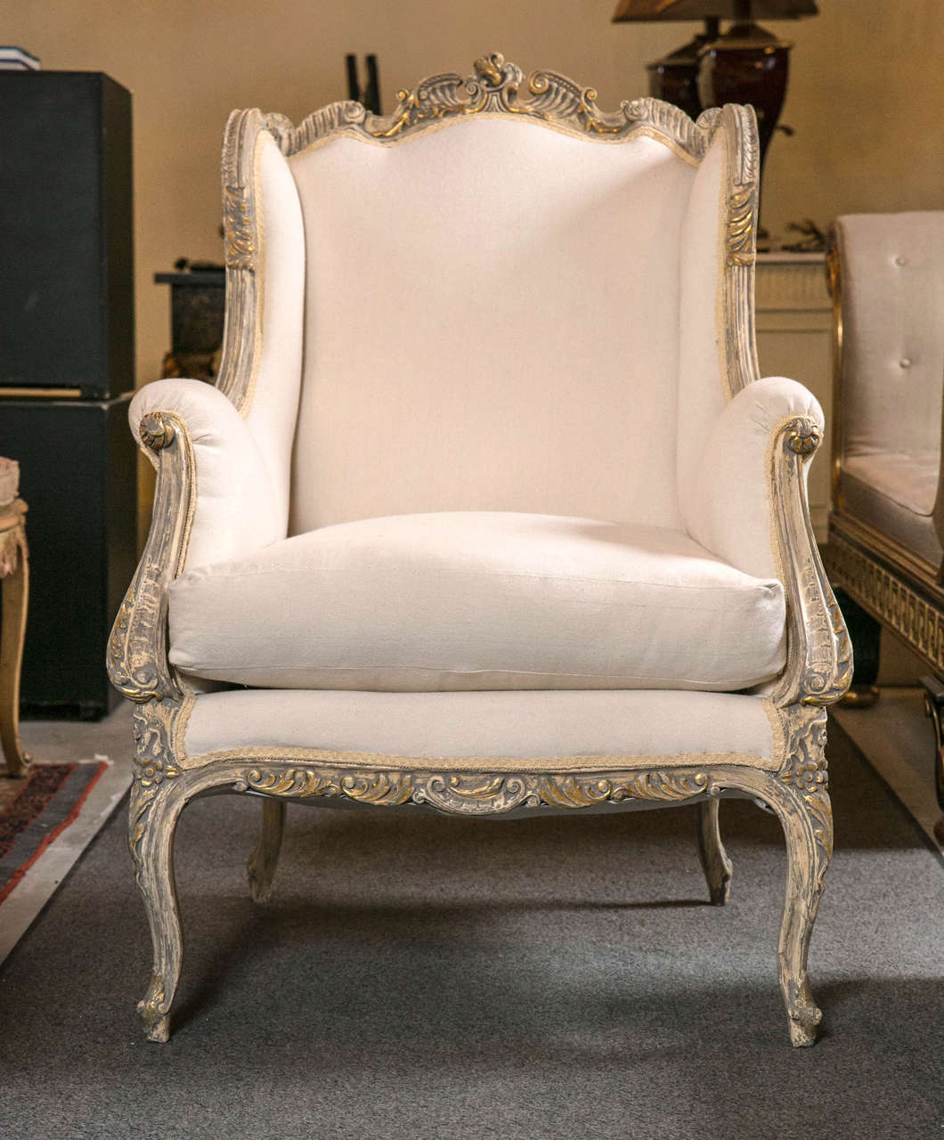 Pair of vintage French Rococo style wingback bergère chairs, circa 1940s, decorated in this gorgeous distressed ivory/grey finish and parcel-gilding, the shell and scroll crest leading to winged sides and scrolled arms, padded back and cushioned