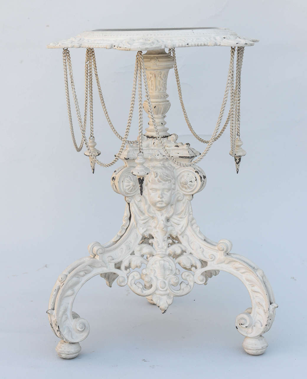 Pedestal or stand, of wrought iron, having distressed painted finish, top inset with round beveled glass, raised on tripartite base with putti heads and draped with metal chain.

Stock ID: D9228
