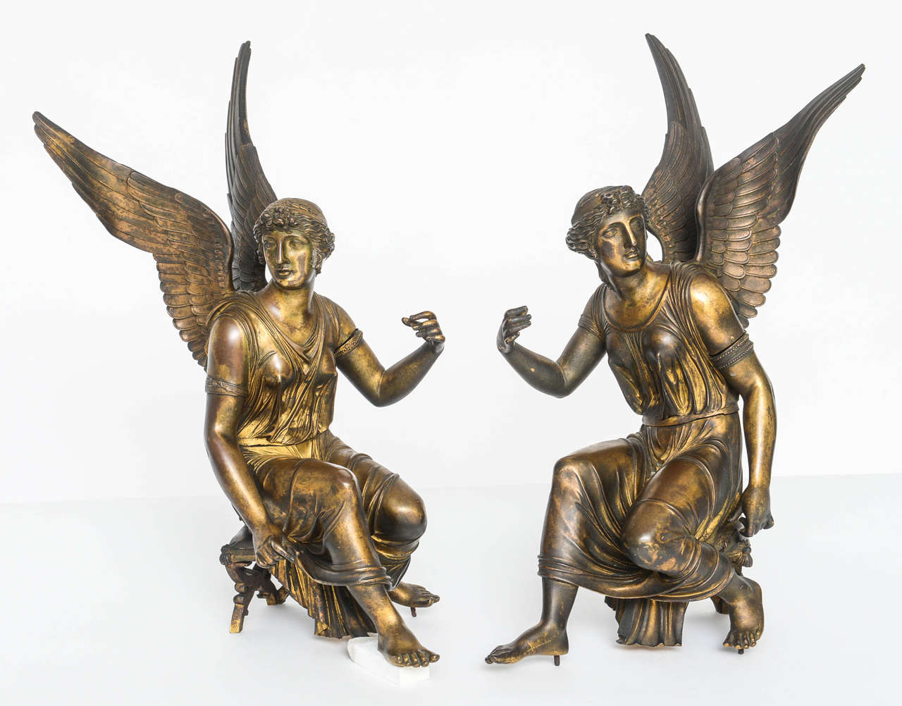 This arrestingly beautiful pair of gilt bronze depictions of Nike the goddess of victory seated on curule stools. They are near mirror images of each other. The arms are outstretched and may have once grasped poles surmounted with laurel wreaths.