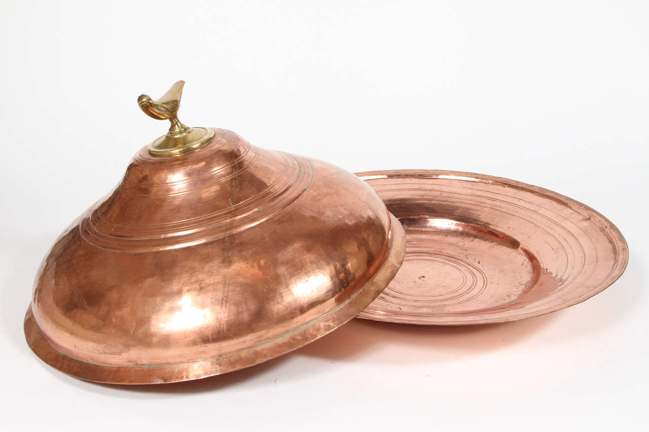 Very nice handcrafted round Moroccan copper tajine serving dish with cover.
The tagine is a copper platter with a cover featuring a brass bird as a handle.
Moroccan tajine with nice patina and a warm glow.


