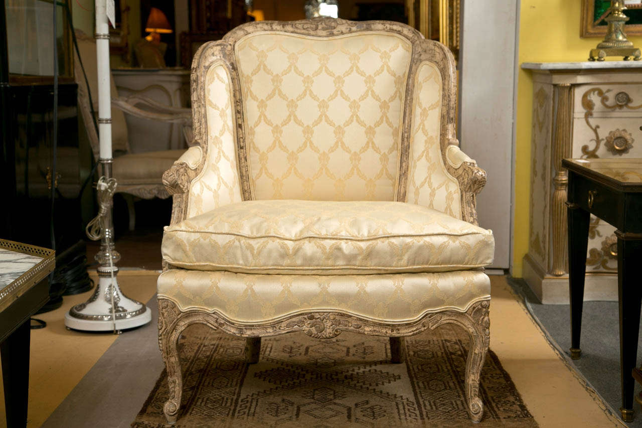 Pair of French Louis XV style winged bergere chairs, circa 1940s, each having distressed frame, the shaped back with downswept arms, padded back and cushioned seat, upholstered in soft-gold sateen fabric, raised on cabriole legs. 

Seat Height: 18