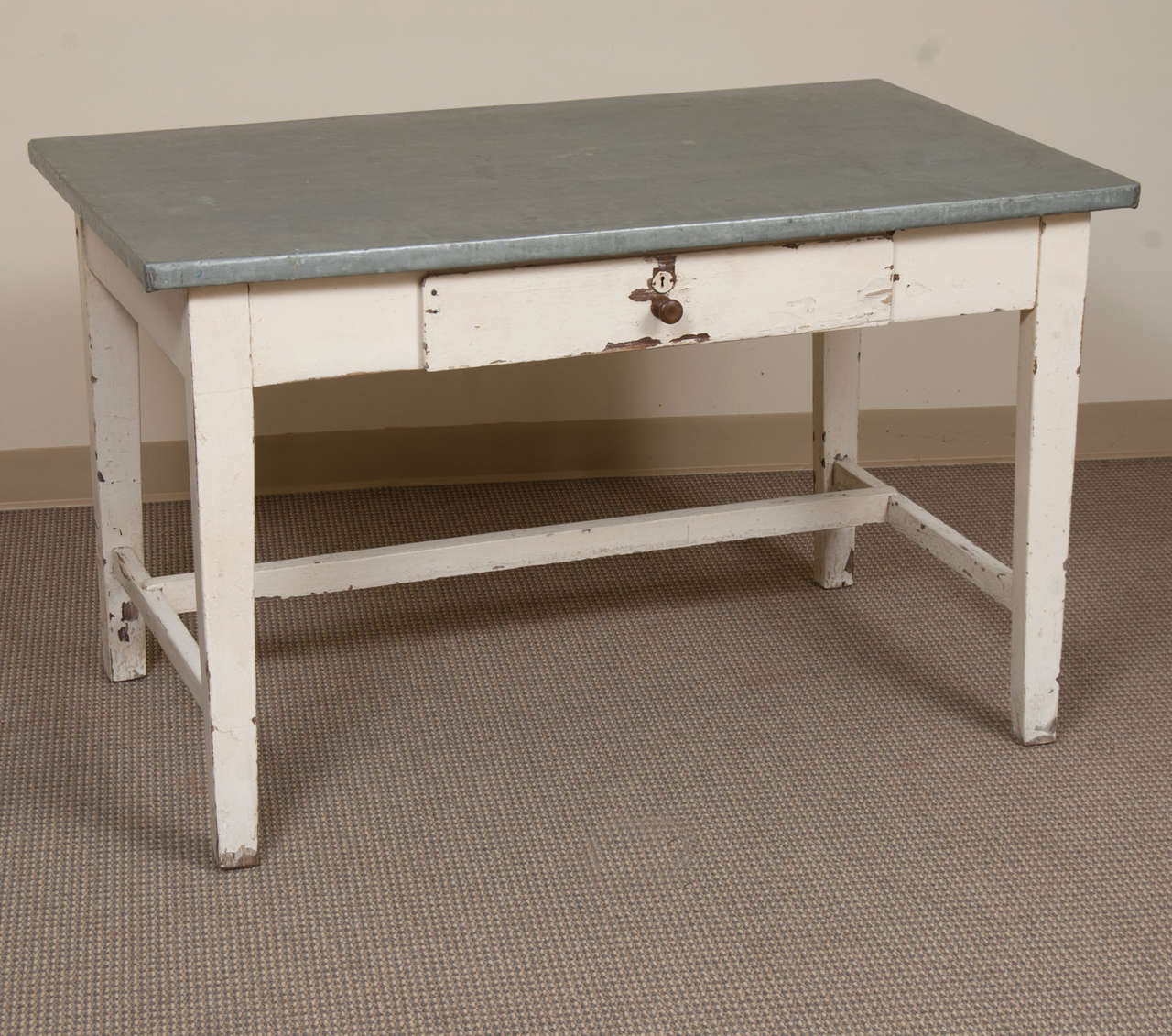 A sturdy and utilitarian pine and oak writing or work table featuring a zinc top in excellent condition, a single central drawer and an off-centre stretcher to the distressed white-painted base.